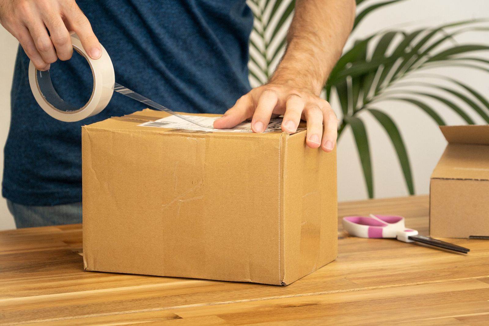 Mãos fechando com fita uma caixa de papelão sobre uma bancada de expedição